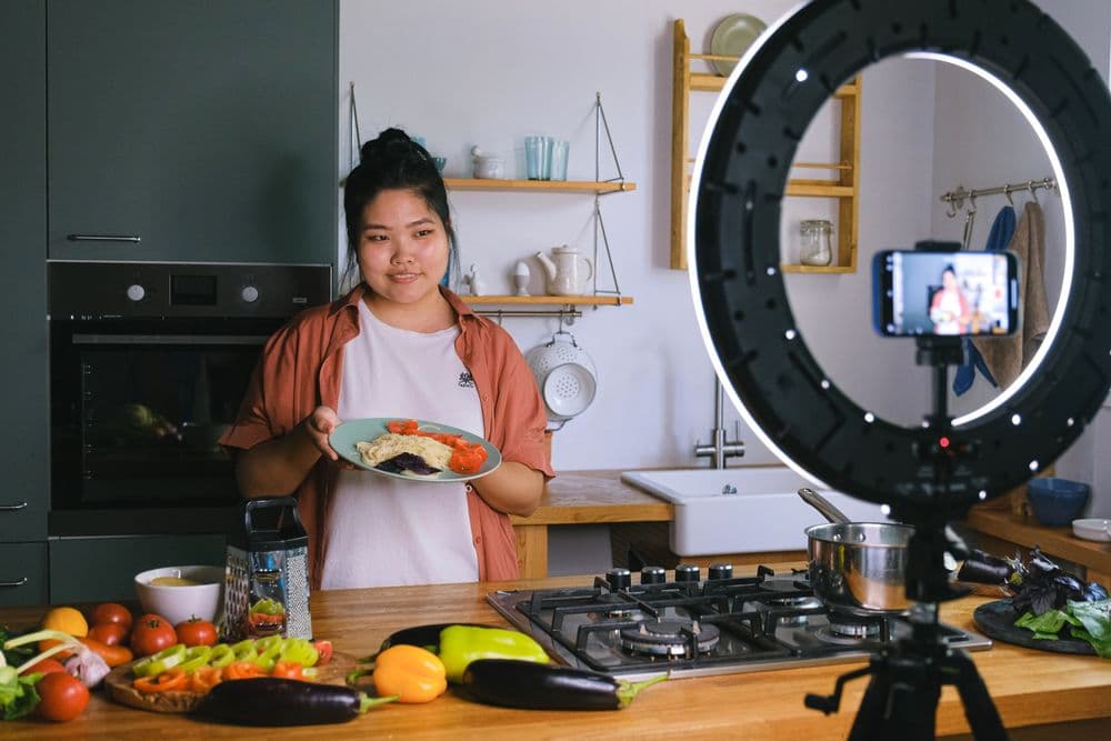 A cook holding a plated dish while a phone on a ring light films the kitchen